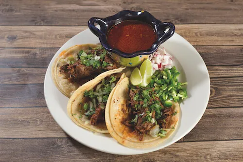 Three tacos with lime and cilantro beside a small bowl of red salsa