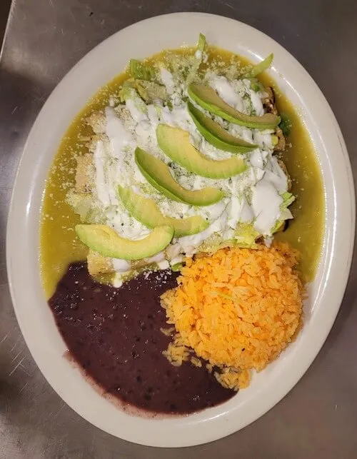 Enchiladas in green sauce topped with avocado and crumbled cheese, with rice and beans
