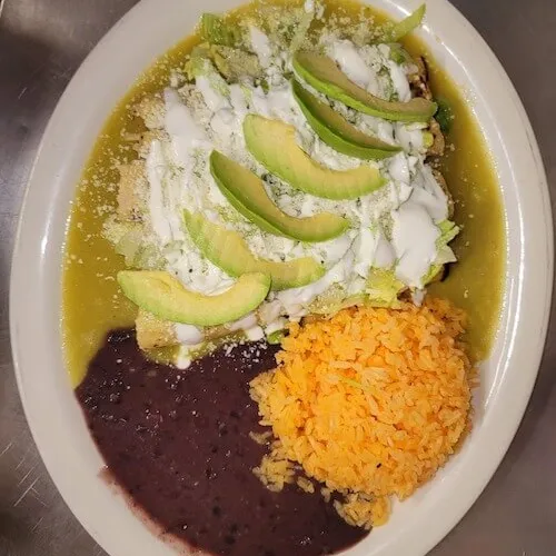 Enchiladas in green sauce topped with avocado and crumbled cheese, with rice and beans